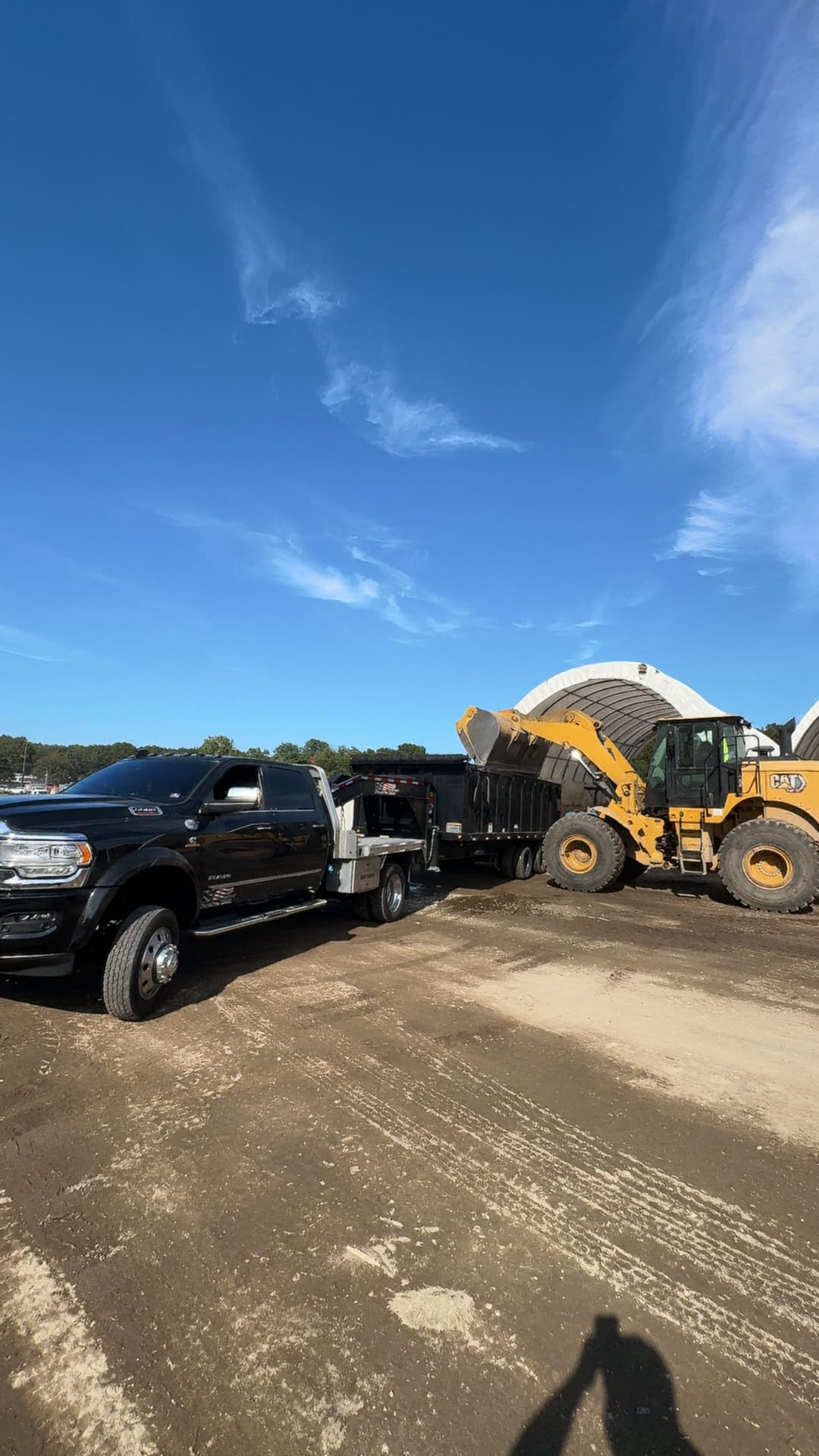 Black pickup truck towing a trailer next to a large yellow front loader on a construction site.