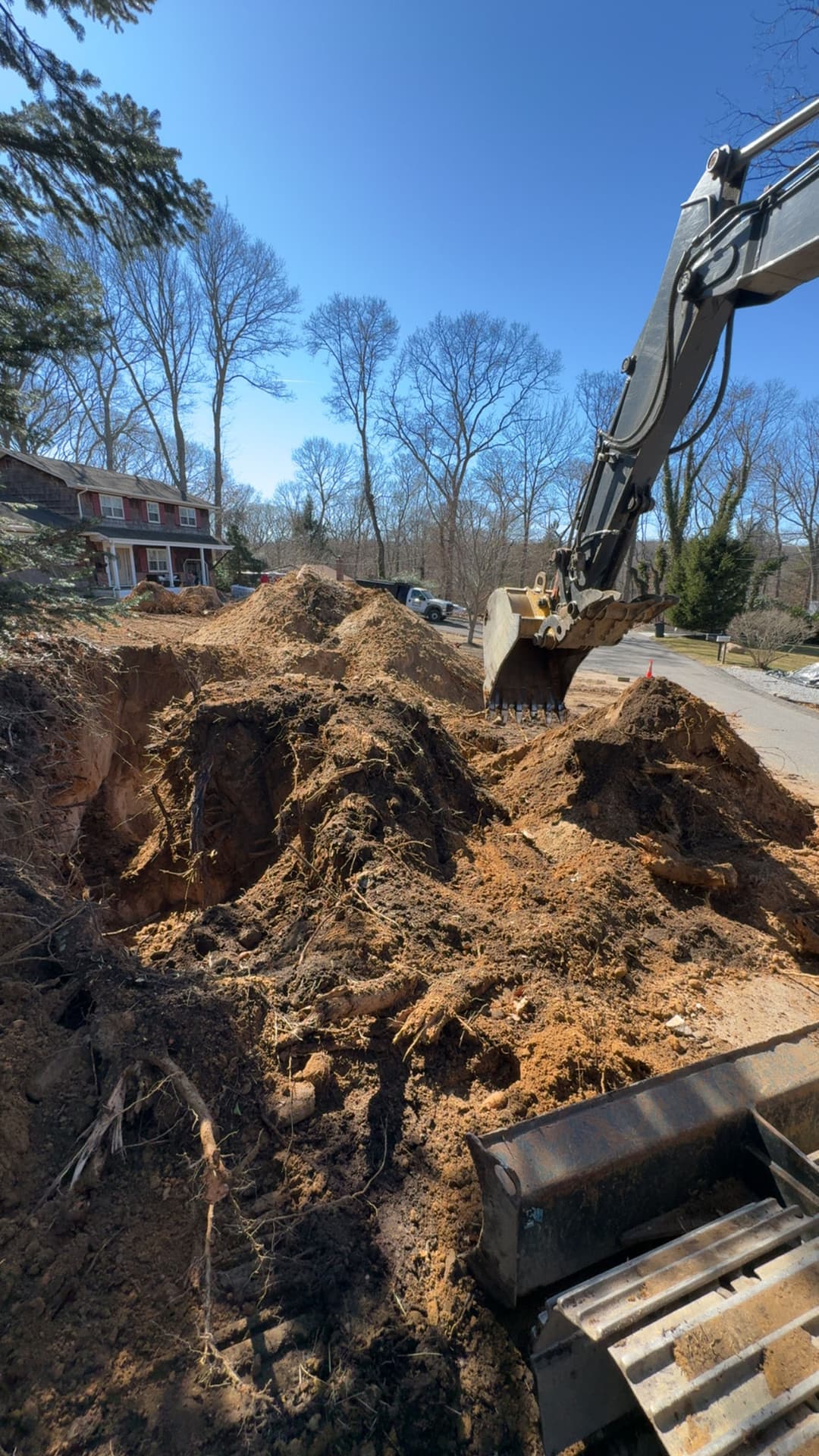 Excavator digging dirt pile on residential street under clear blue sky.