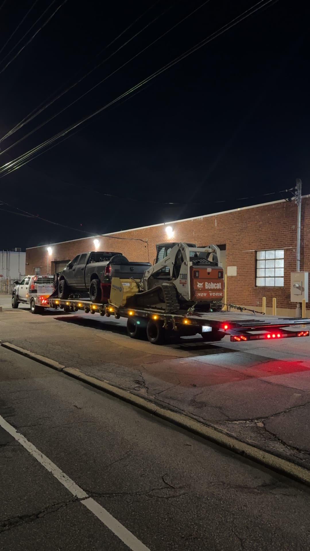 Tow truck transporting a damaged vehicle and excavator at night. Urban setting with industrial backdrop.