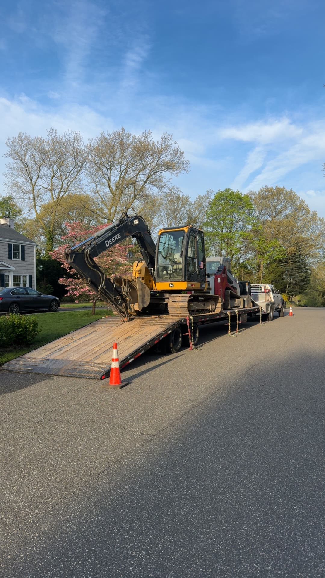 Excavator on flatbed truck parked on residential street with trees and clear sky.