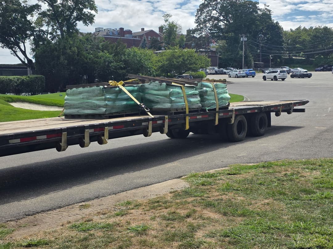 Flatbed truck unloading wrapped materials at a construction site with parked vehicles in background.