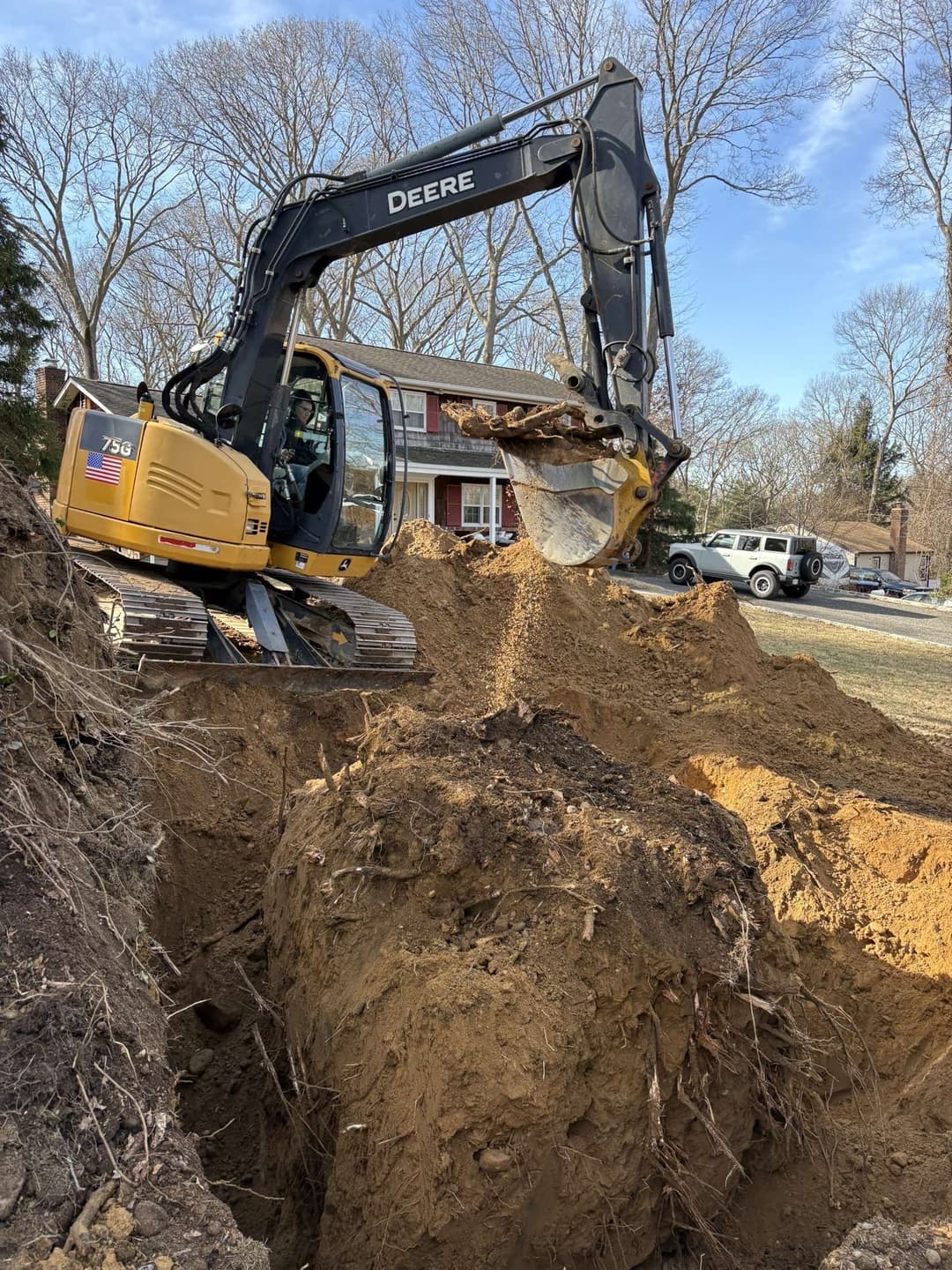 Excavator digging a large trench in a residential yard with a house and truck in the background.