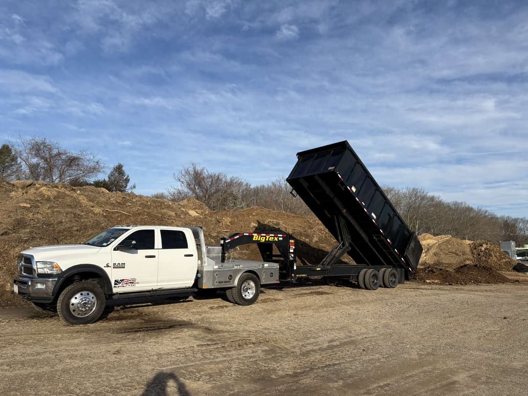 White pickup truck with a black dump trailer unloading dirt at a construction site.