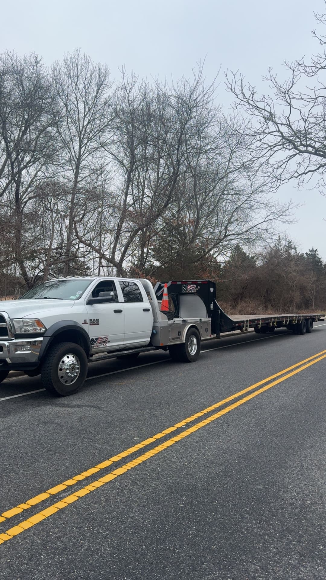 White pickup truck with flatbed trailer parked on a rural road surrounded by trees.