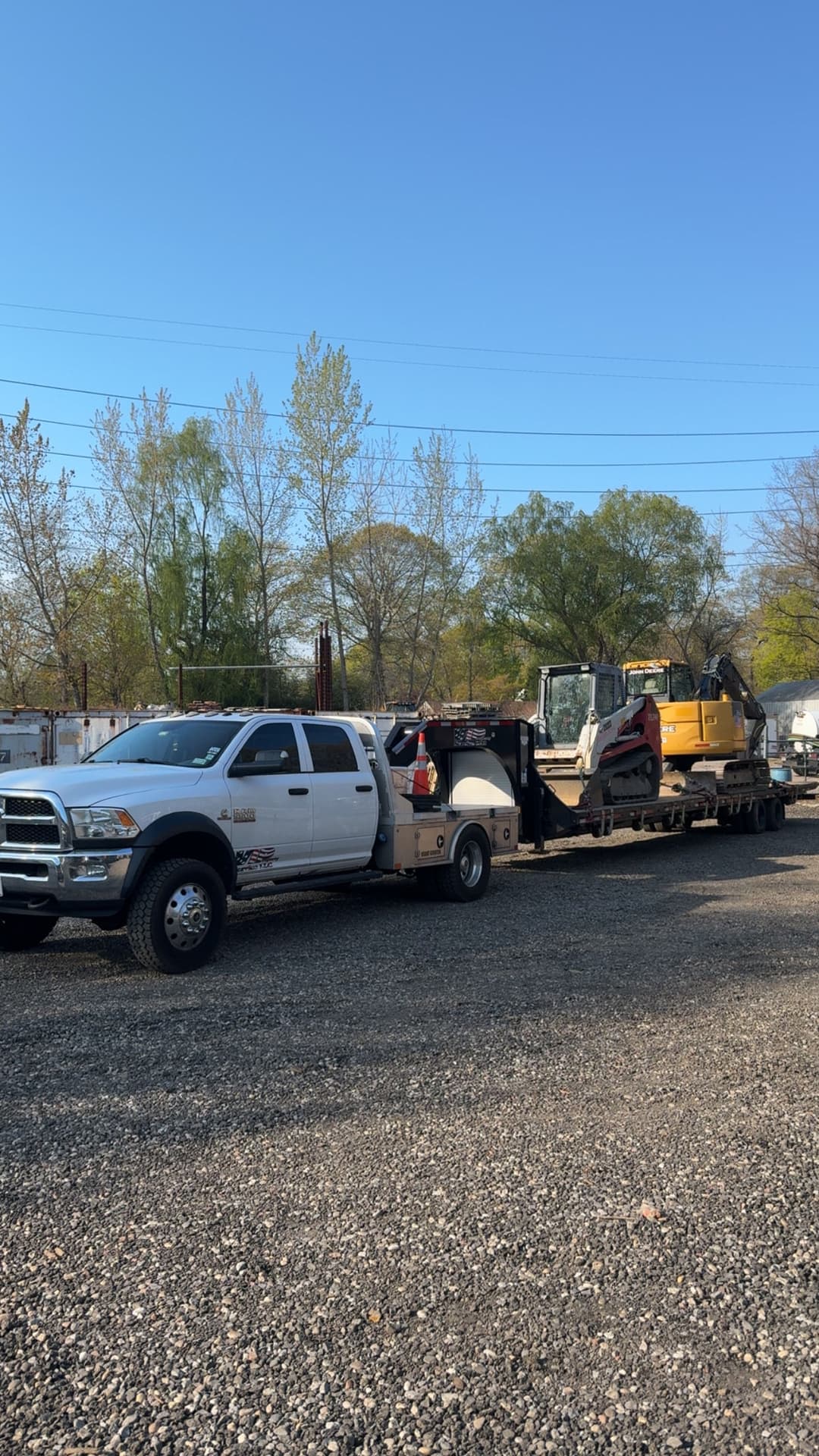 Pickup truck towing heavy machinery on a flatbed trailer in a construction yard.