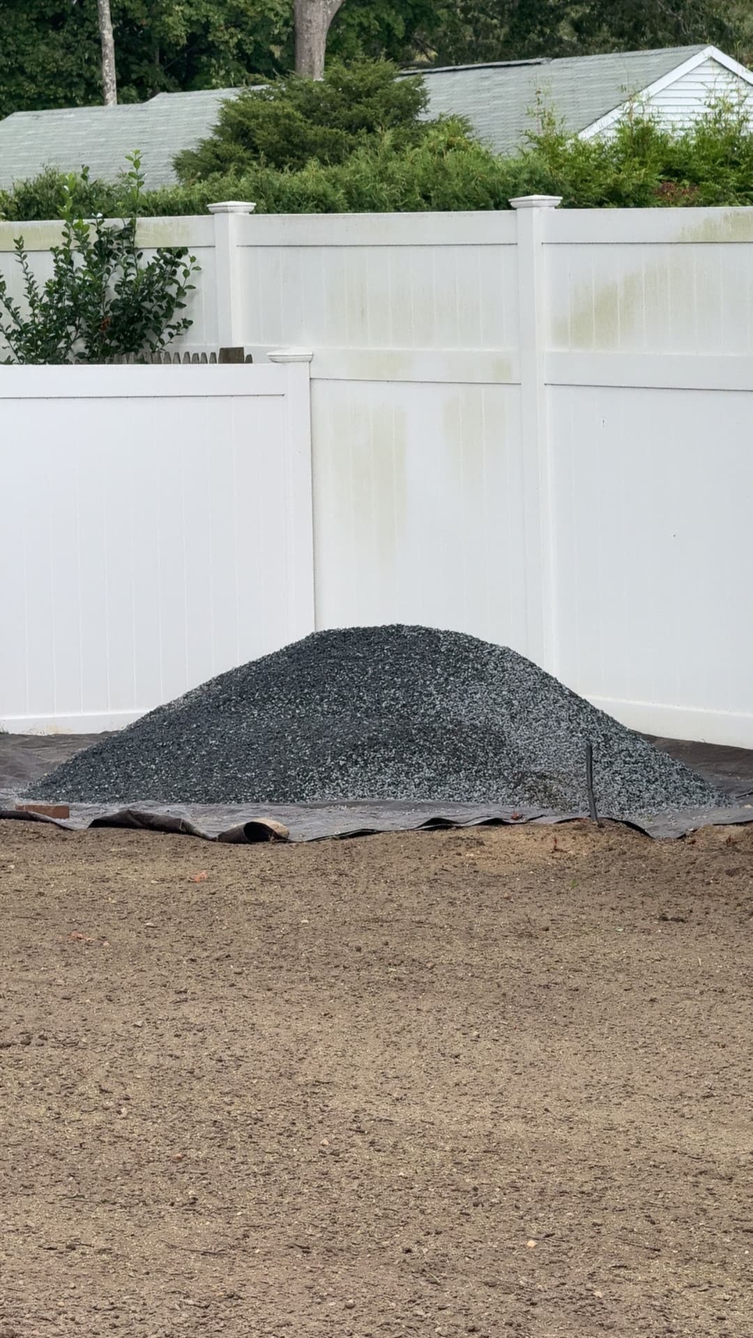 Piled gravel on a tarpaulin in a yard with a white fence in the background.