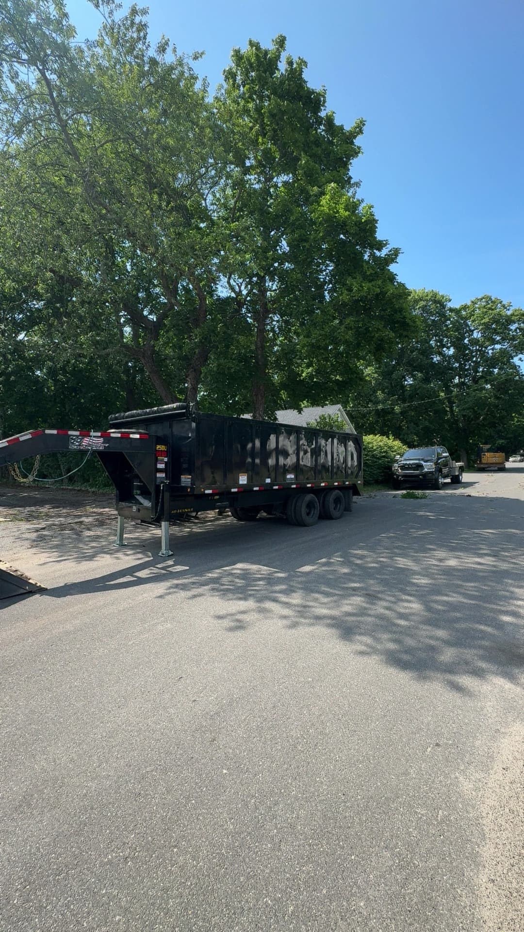 Black dump trailer parked on a sunny road, surrounded by greenery and trees.