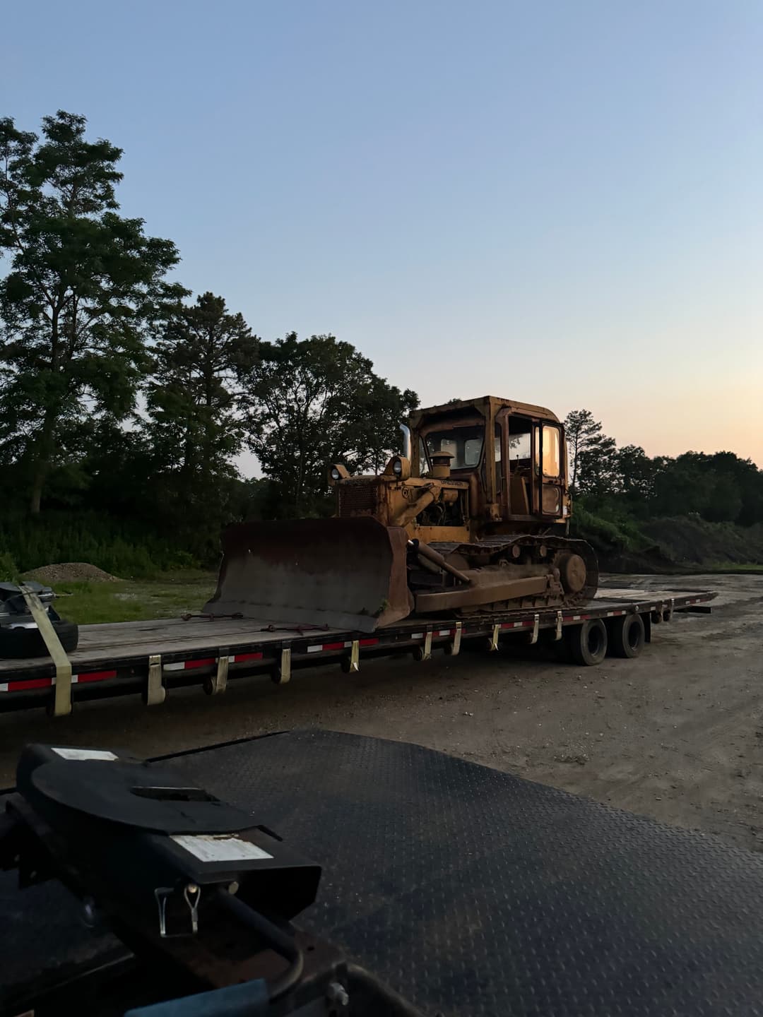 Bulldozer loaded on flatbed trailer at dusk surrounded by trees and open land.