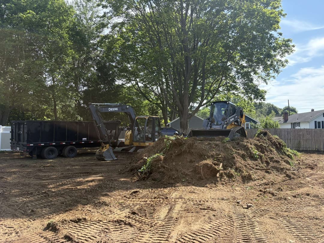 Excavators and dump truck on a residential construction site with dirt pile and trees.