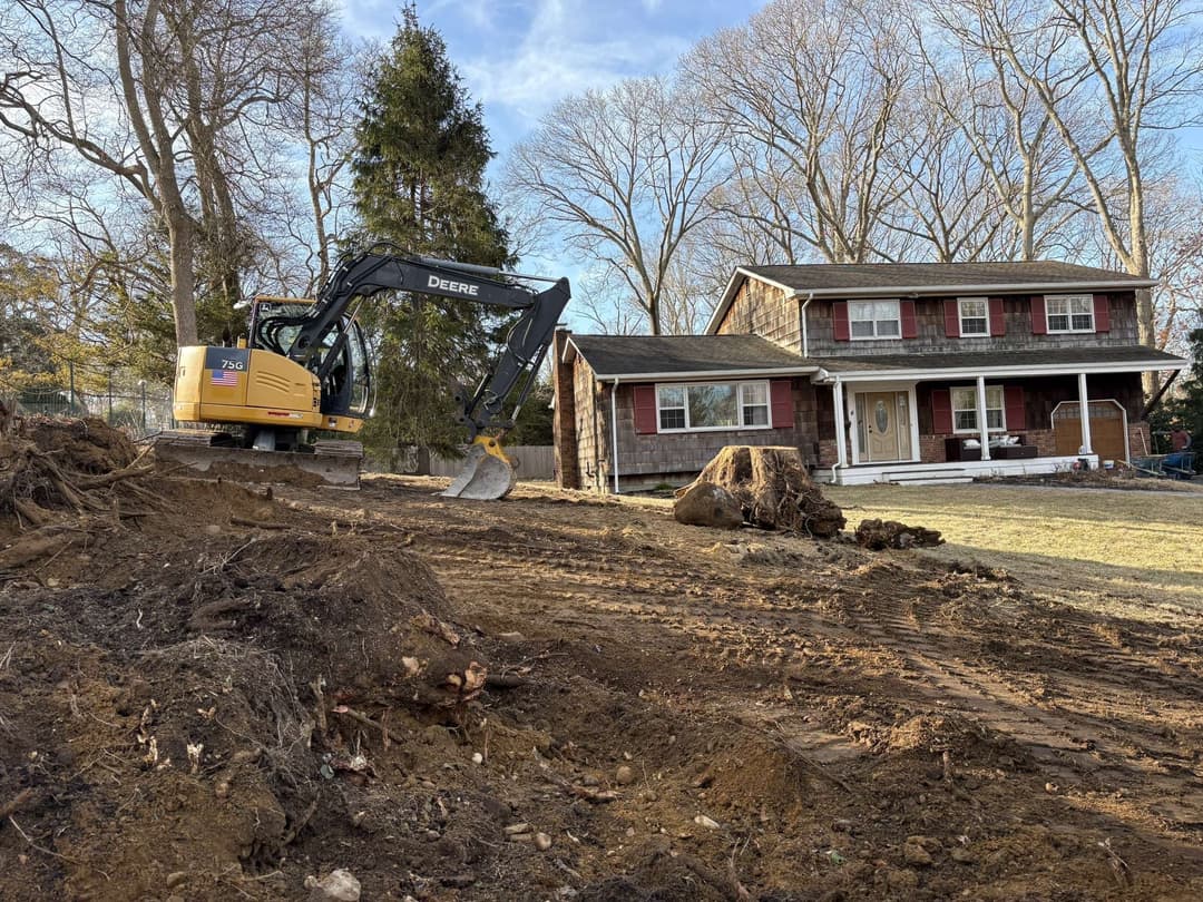 Excavator clearing land for landscaping near a residential house with trees.