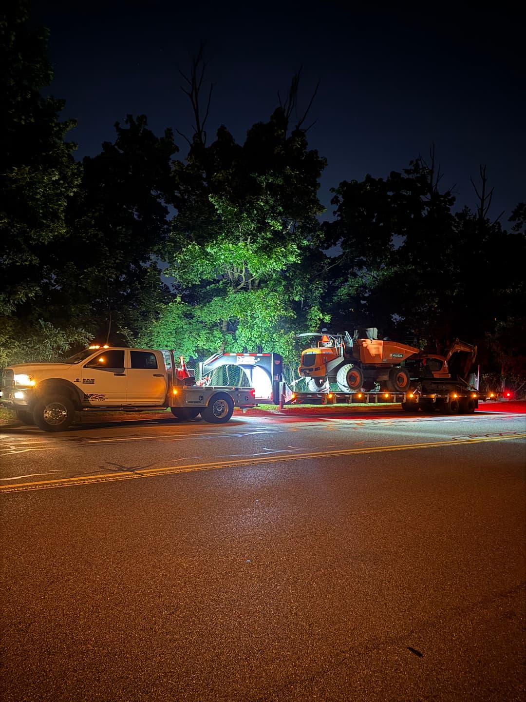 Tow truck transporting construction equipment at night with illuminated surroundings.