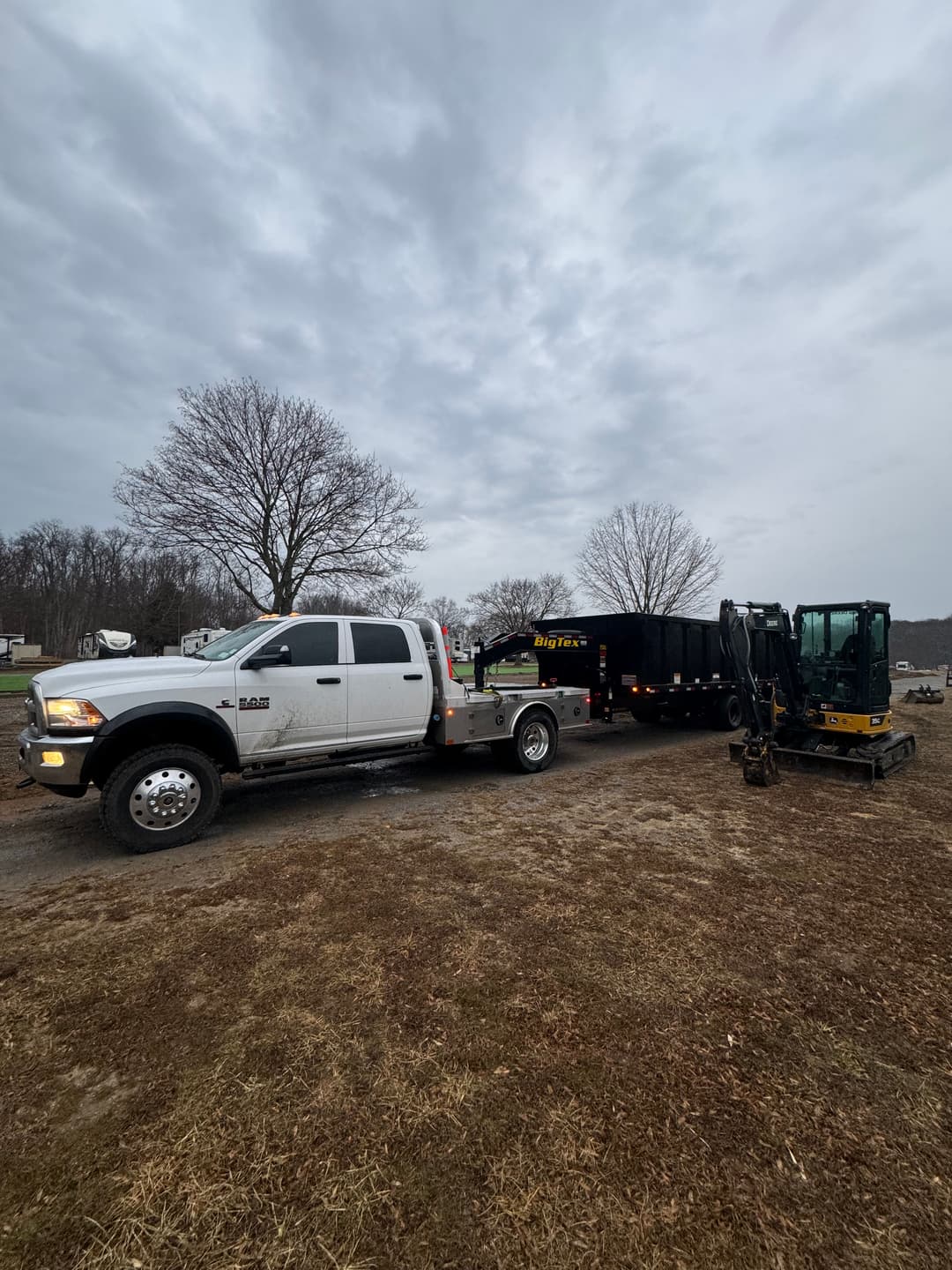 White pickup truck towing a trailer with construction equipment under a cloudy sky.