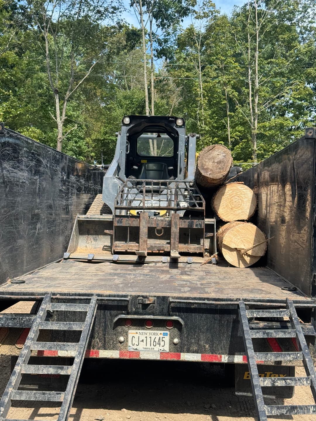 Skid steer loader on trailer with logs, surrounded by trees in a wooded area.