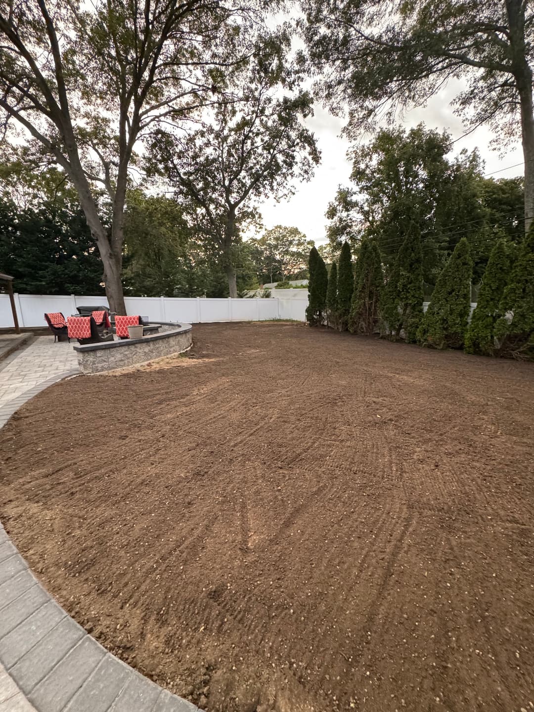 Empty backyard with freshly tilled soil, lined by evergreen trees and a patio with chairs.