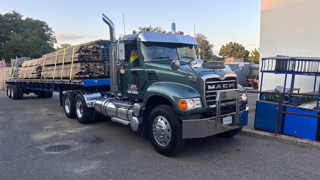 Mack truck with flatbed trailer loaded with lumber parked outside a building.