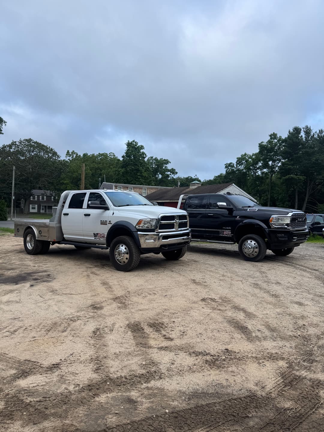 Two heavy-duty trucks parked on a dirt lot surrounded by trees and buildings.