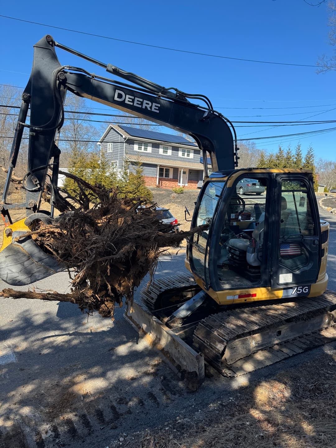 Excavator lifting large tree stump on a residential street with houses in the background.