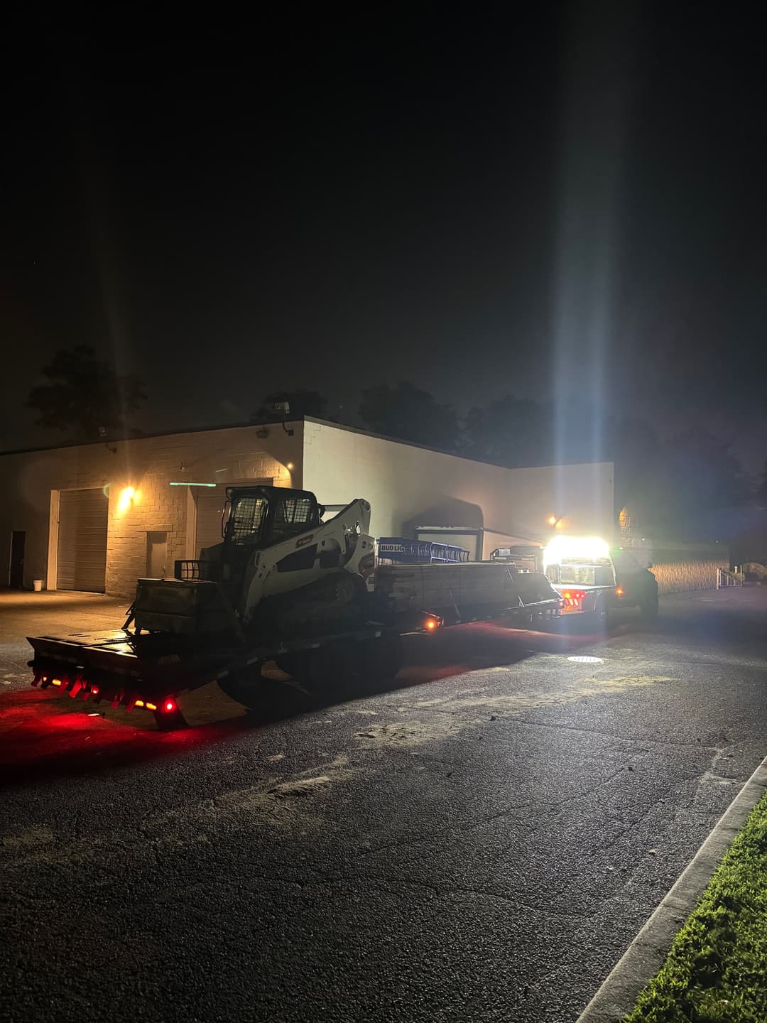 Bobcat with a trailer at night, illuminated by work lights in a parking lot.