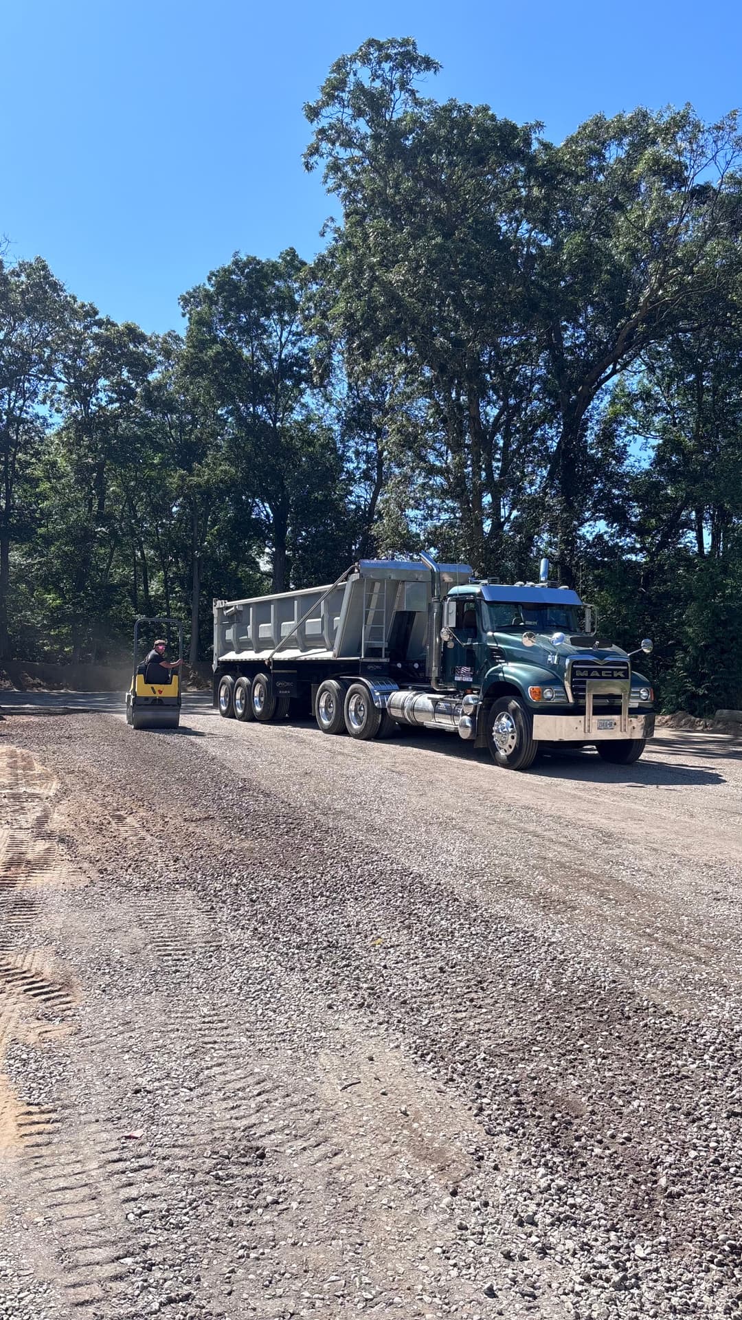 Construction site with a large truck and roller, surrounded by trees under a clear blue sky.