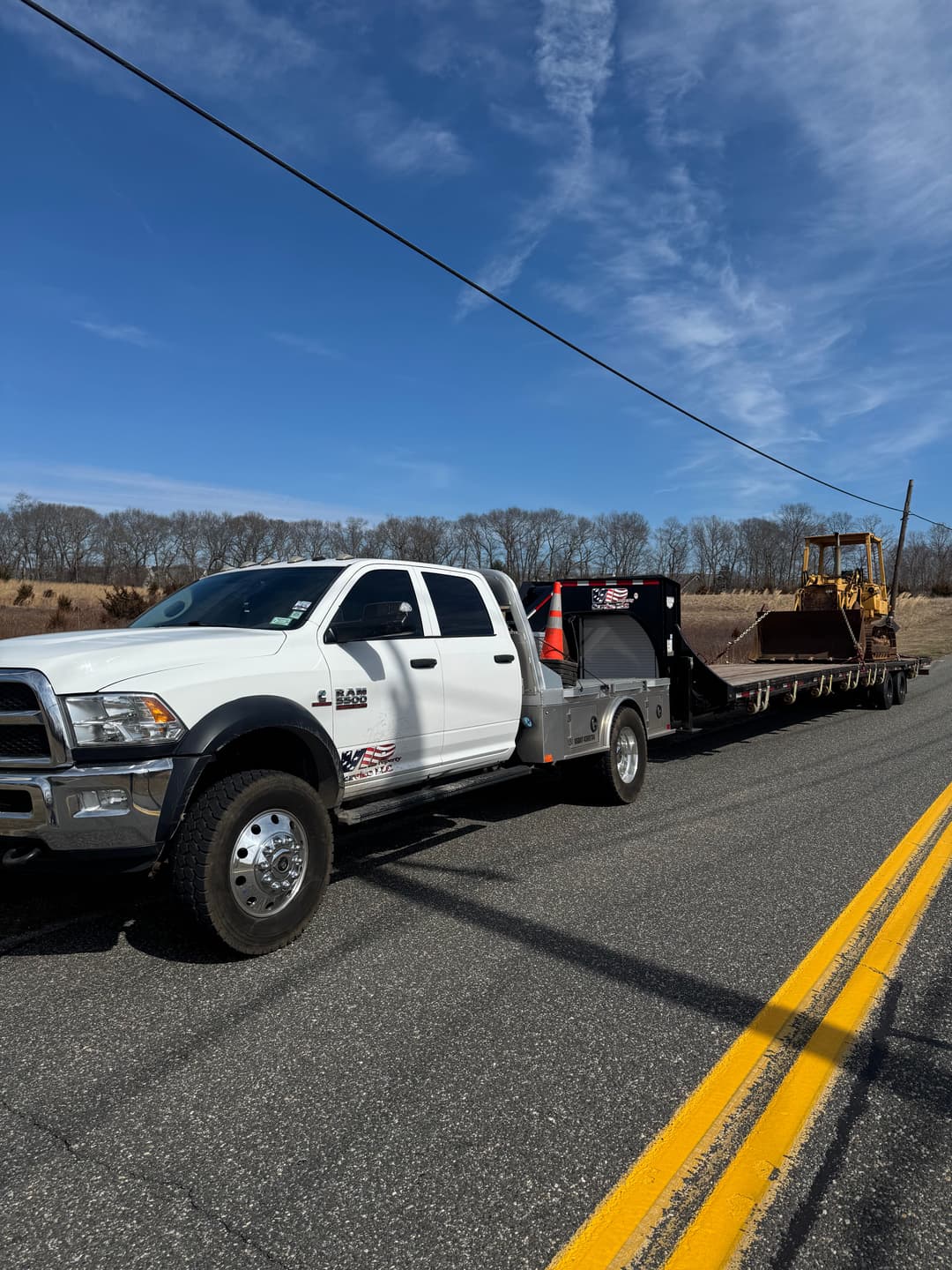 White pickup truck towing heavy machinery on a rural road with blue skies.