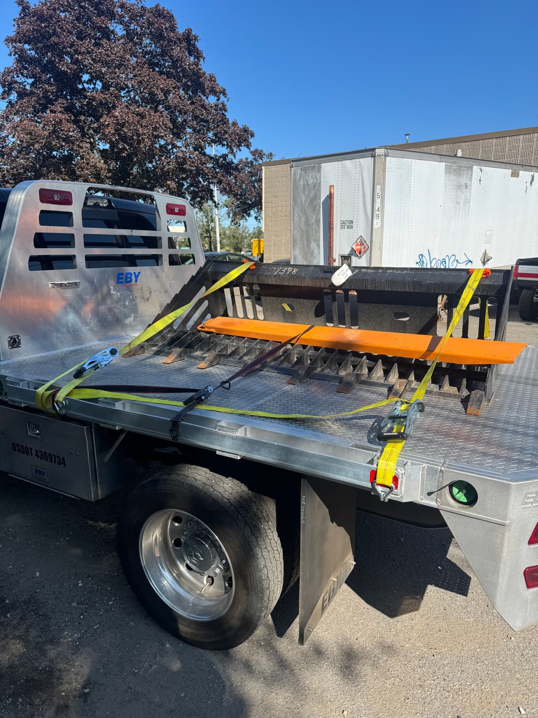 Flatbed truck transporting a heavy steel attachment secured with straps against a clear blue sky.