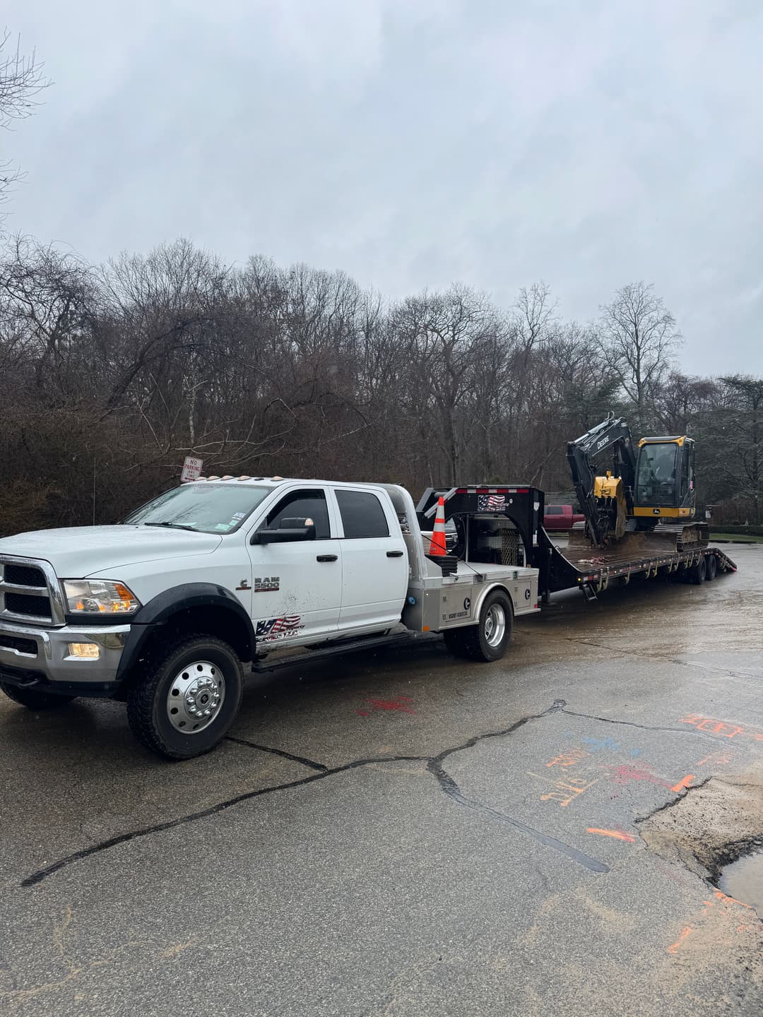 White truck towing a construction vehicle on a flatbed trailer in a rainy setting.