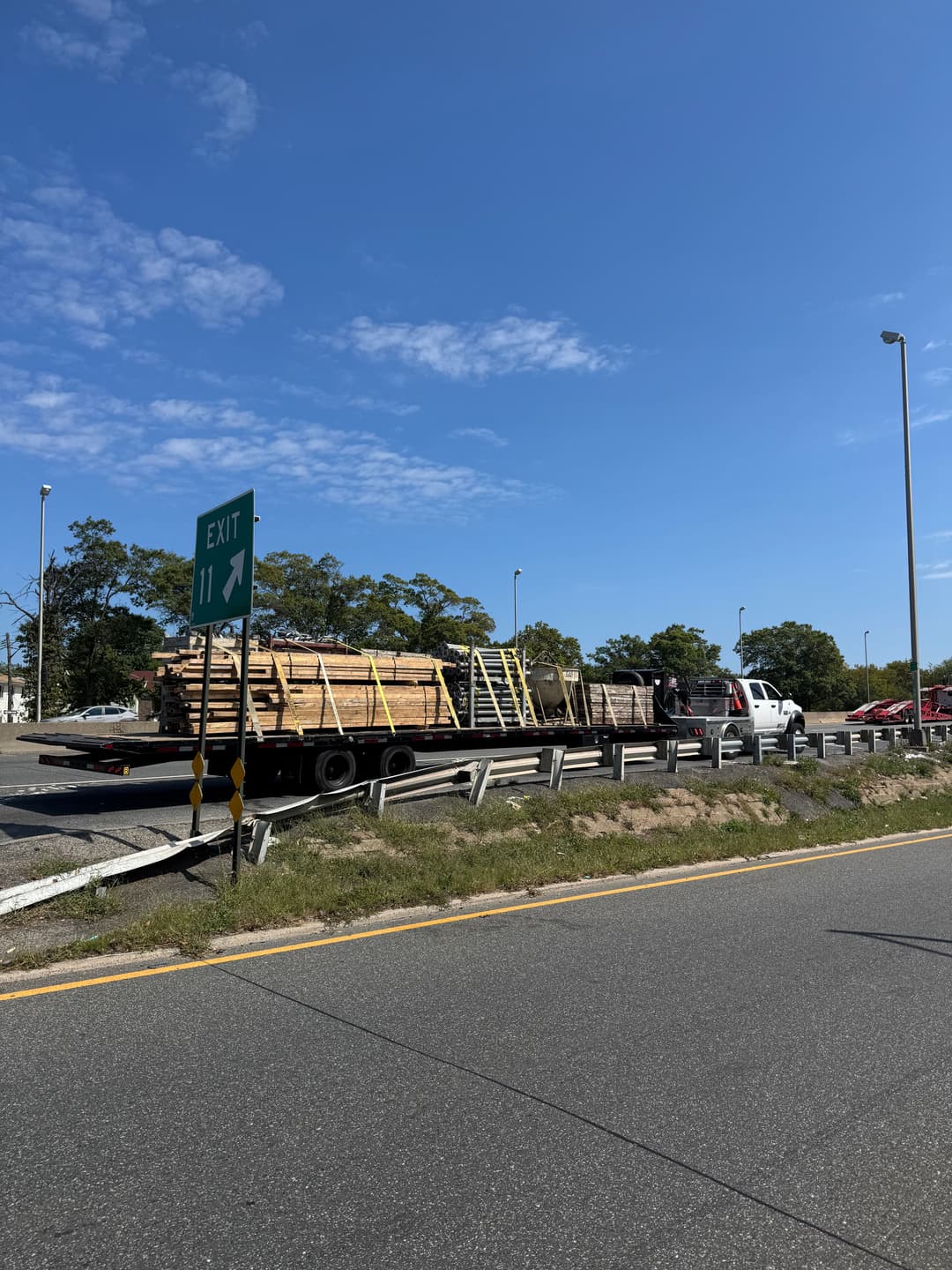 Flatbed truck loaded with lumber on the highway, exit sign visible in the background.