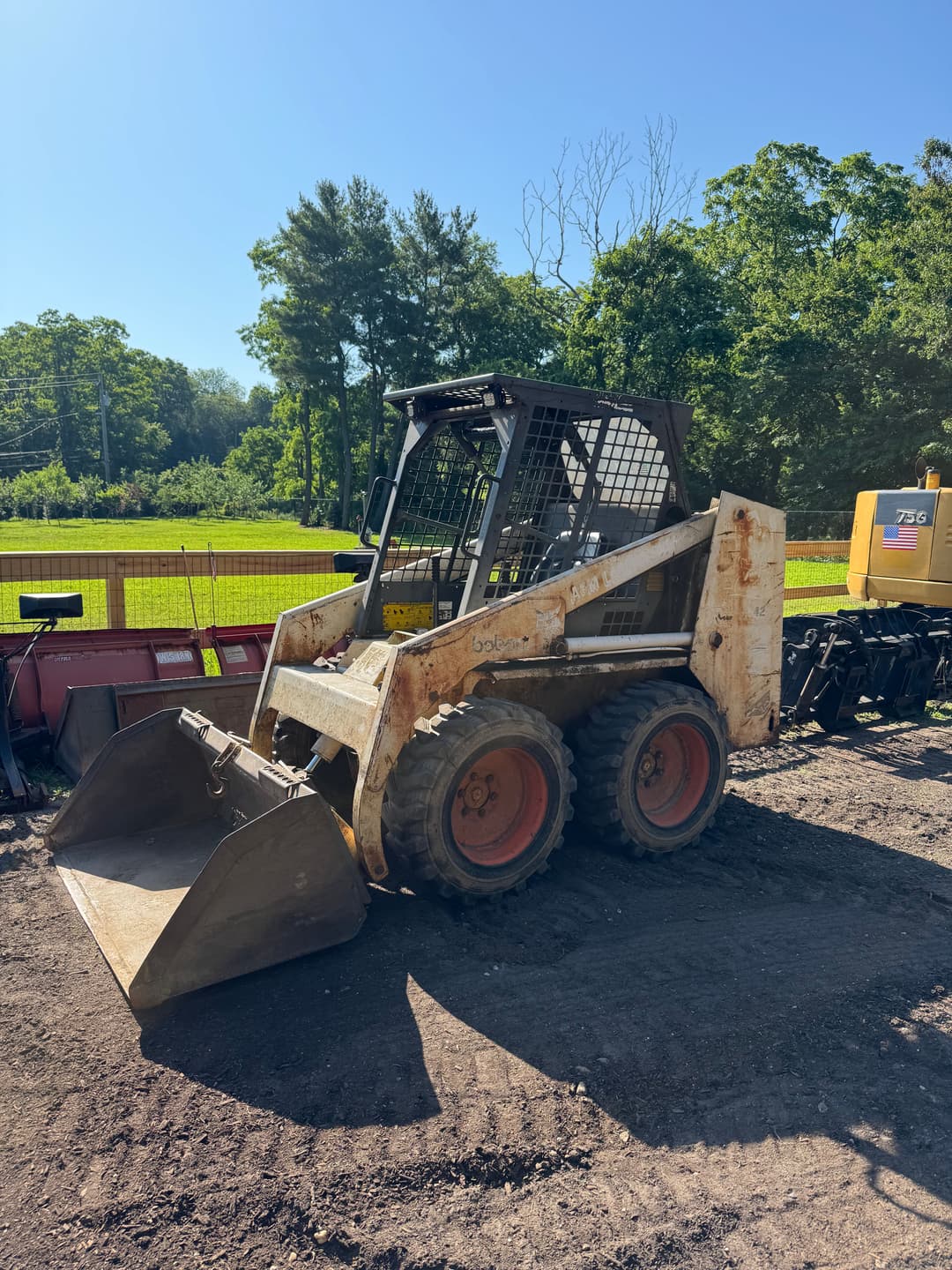 Skid steer loader parked on dirt near construction equipment and greenery.