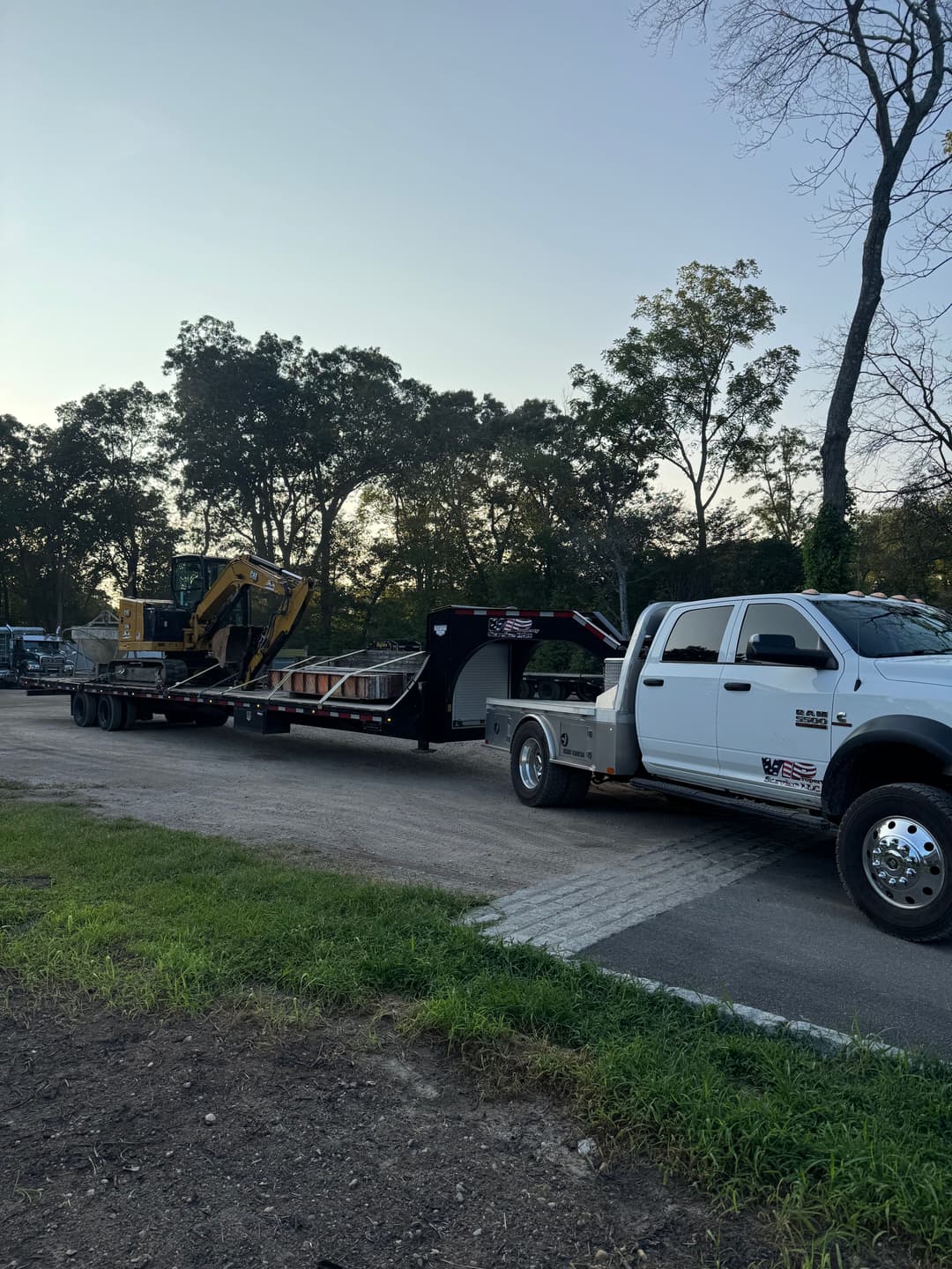 White truck towing a flatbed trailer with heavy machinery in a rural setting at sunset.