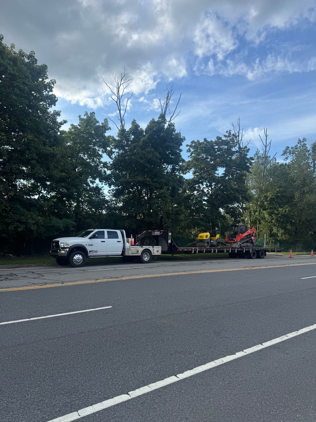 White pickup truck with trailer carrying yellow excavator on a roadside scene.