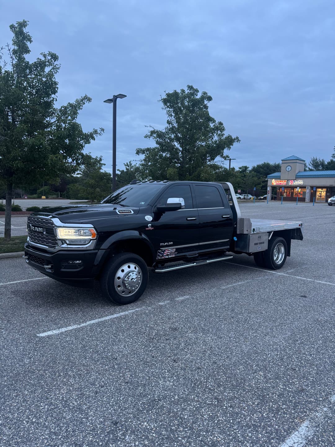 Black RAM truck parked in a lot with a flatbed, surrounded by trees and a cloudy sky.