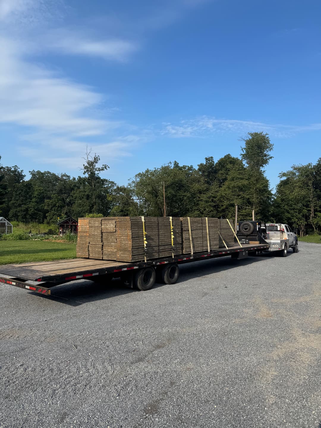 Flatbed truck transporting stacked wooden planks on a gravel lot with green trees in background.