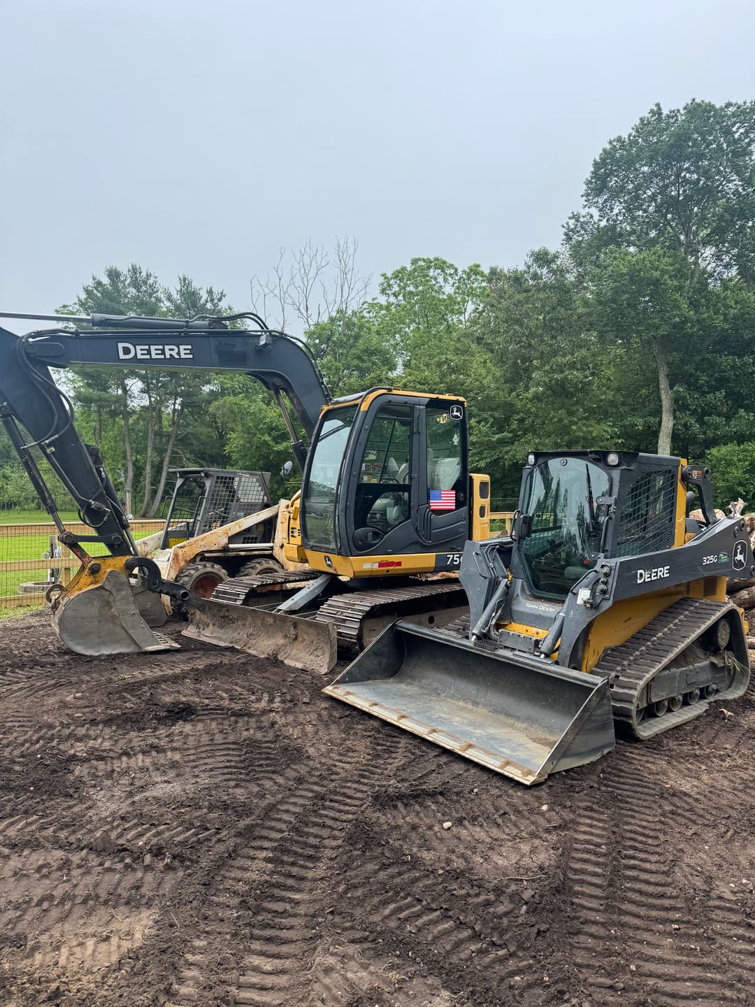 Two John Deere excavators on a construction site with muddy ground and trees in the background.