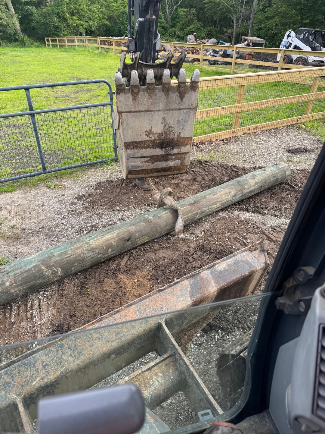 Excavator grasping a log on a construction site with fencing and machinery in the background.