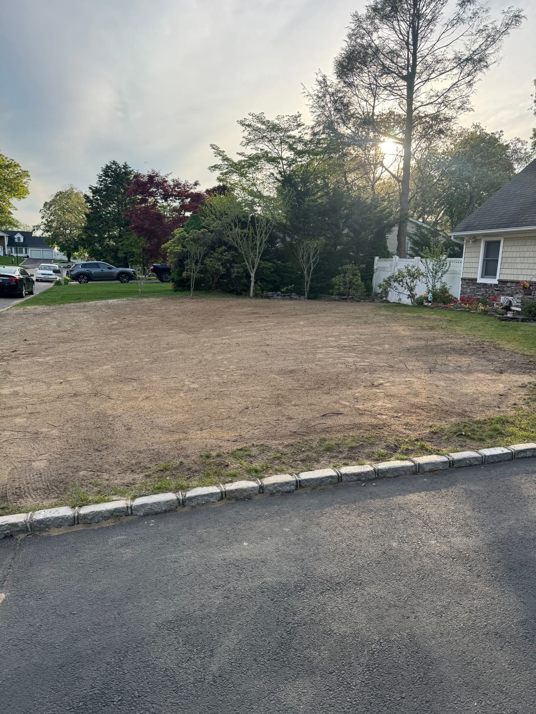 Empty dirt lot with gravel border, surrounded by trees and a residential area, under a sunny sky.
