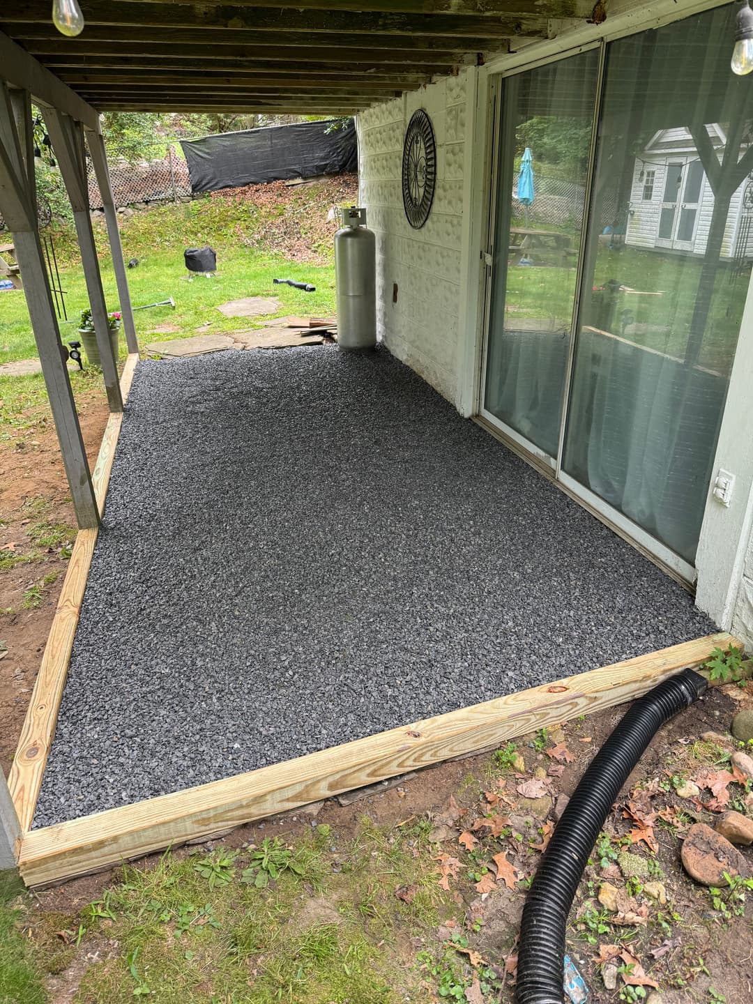Gravel patio installation under a covered porch with wooden frame and drainage hose visible.
