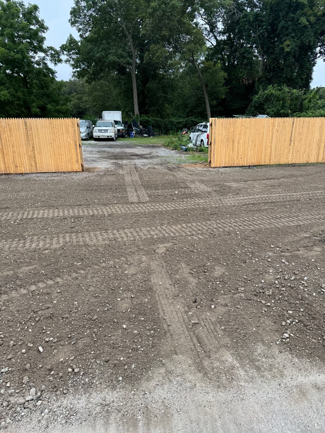 Gravel entrance with tire tracks leading to a wooded area and parked vehicles behind a wooden fence.