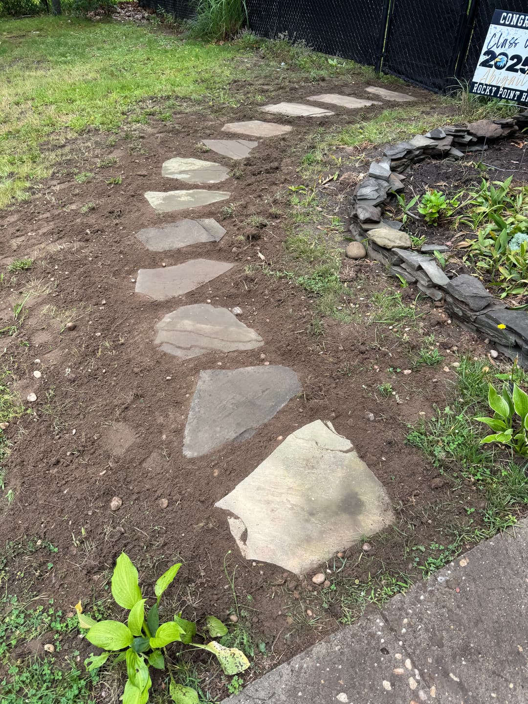 Stone pathway leading through a garden, bordered by soil and plants, with a decorative sign.