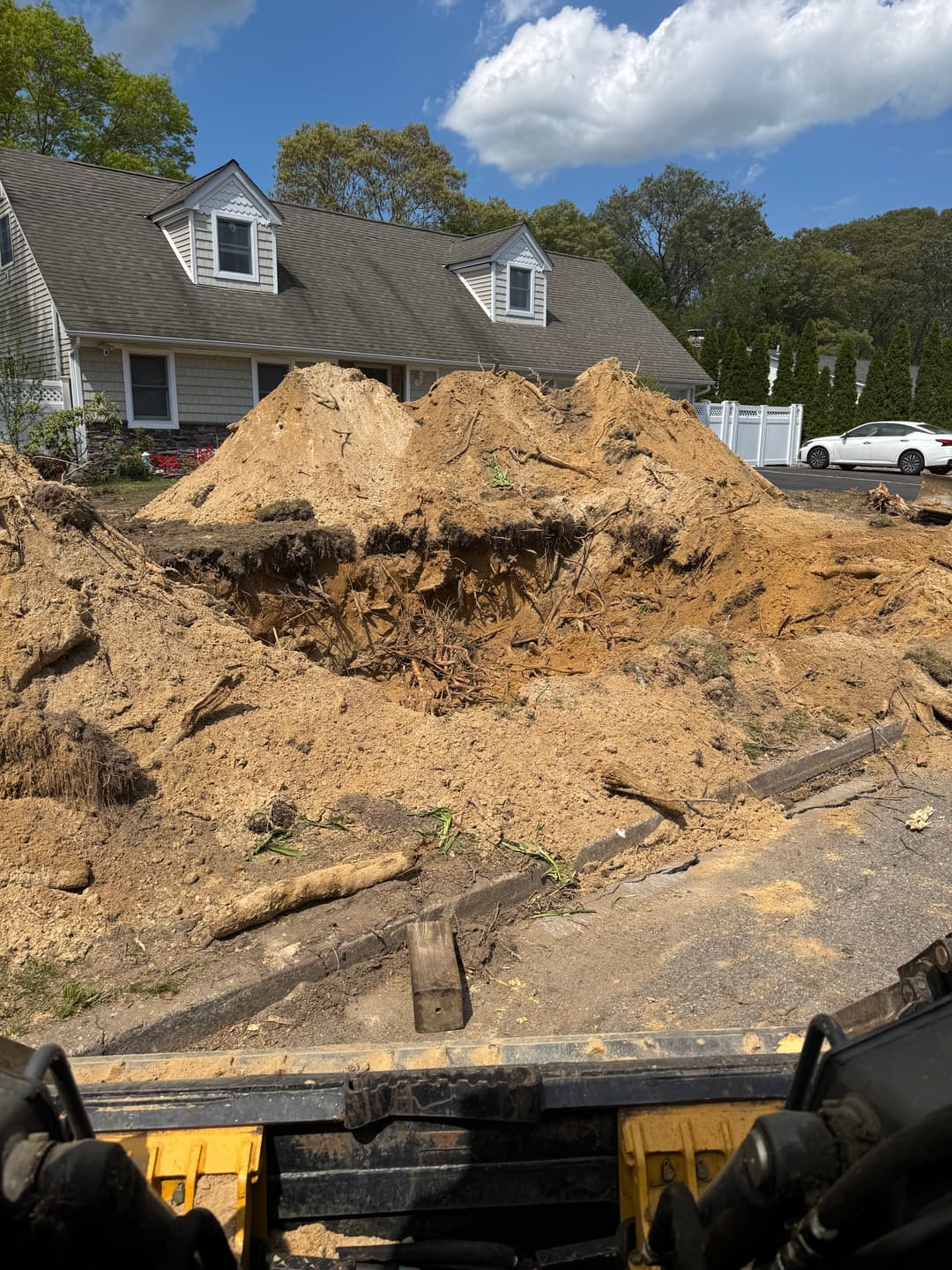 Excavation site with large dirt mound and house in background under clear blue sky.
