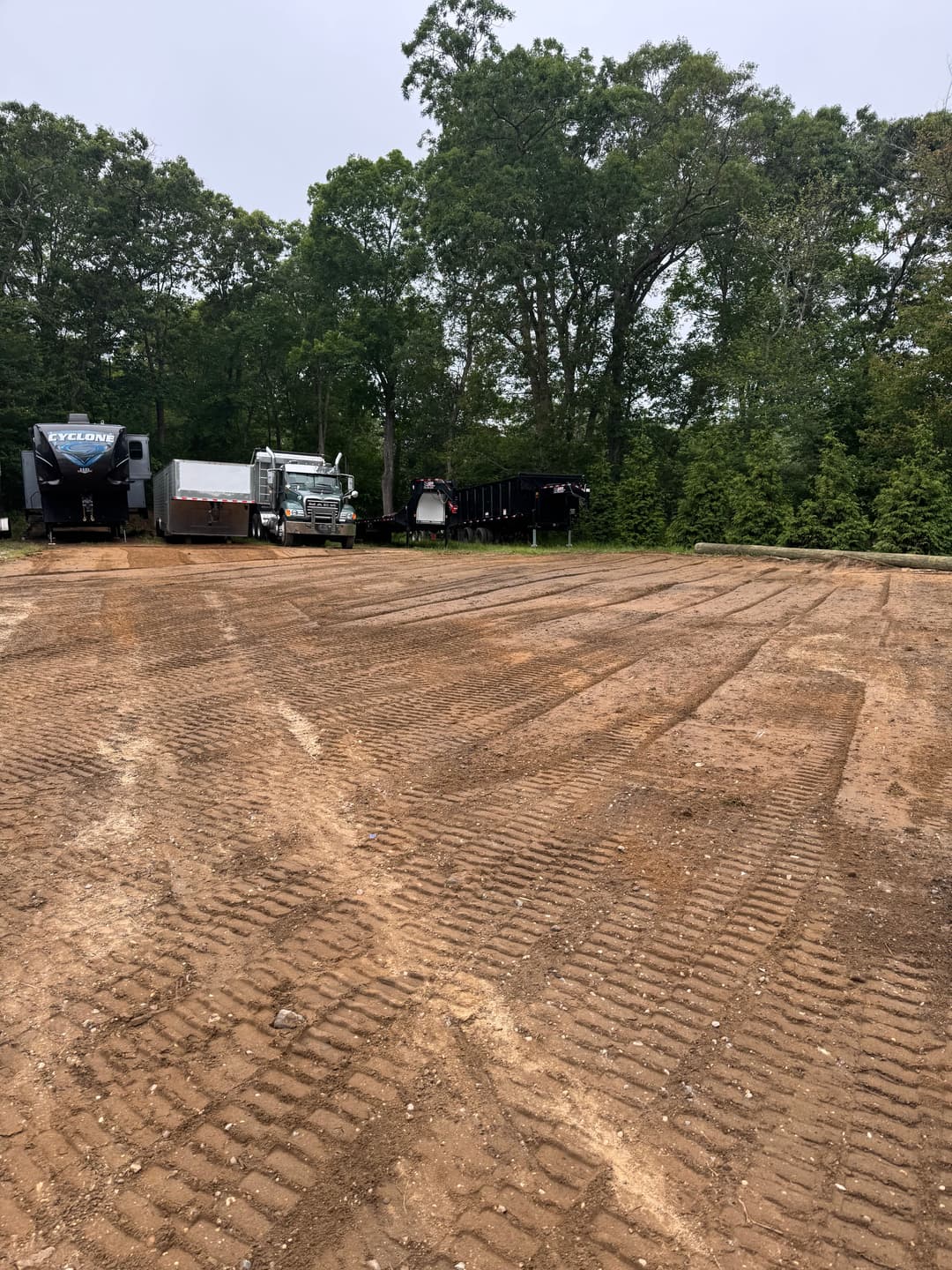 Trucks parked on a gravel lot surrounded by trees, featuring tire tracks on the ground.