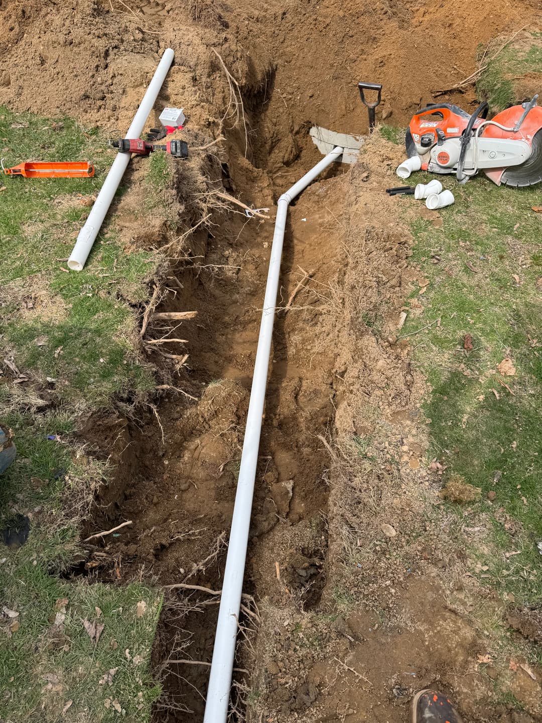 PVC pipe installation in a trench with tools and equipment on a grassy area.
