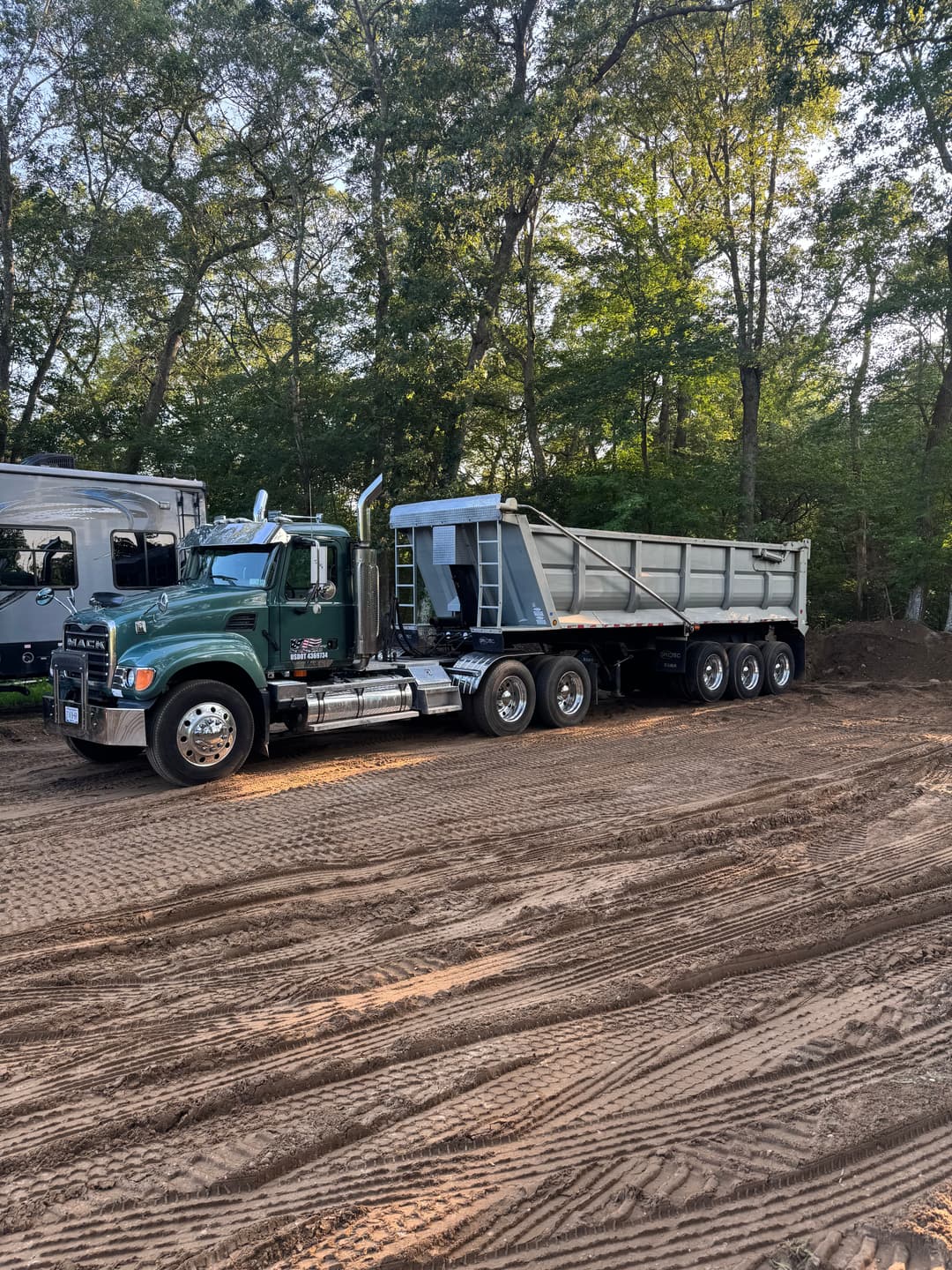 Mack truck with a dump trailer parked on sandy ground surrounded by trees.