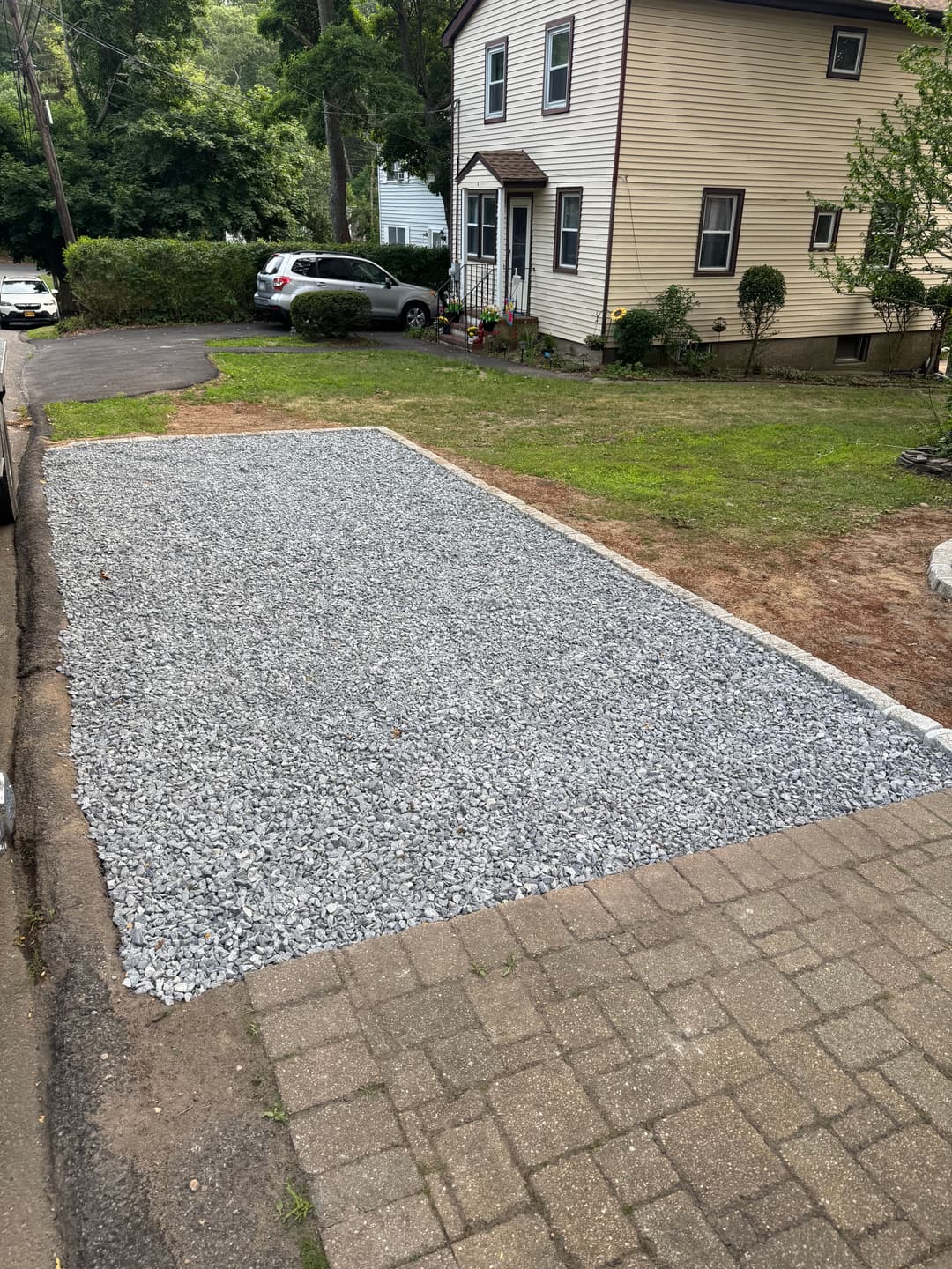 Gravel driveway installation beside a residential home with landscaped yard and parked cars.
