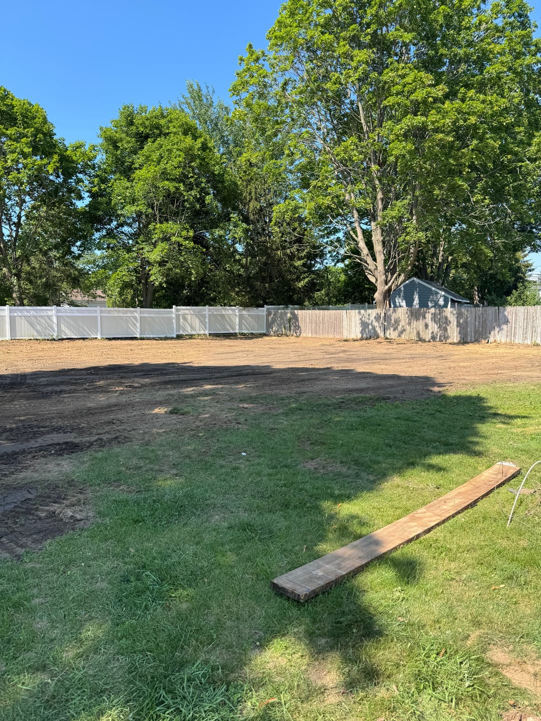 Cleared backyard with lush grass, trees, and a wooden plank, ready for landscaping.
