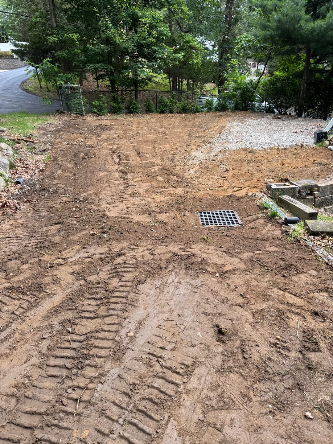 Cleared gravel area with tire tracks, drainage grate, and surrounding trees in a residential setting.