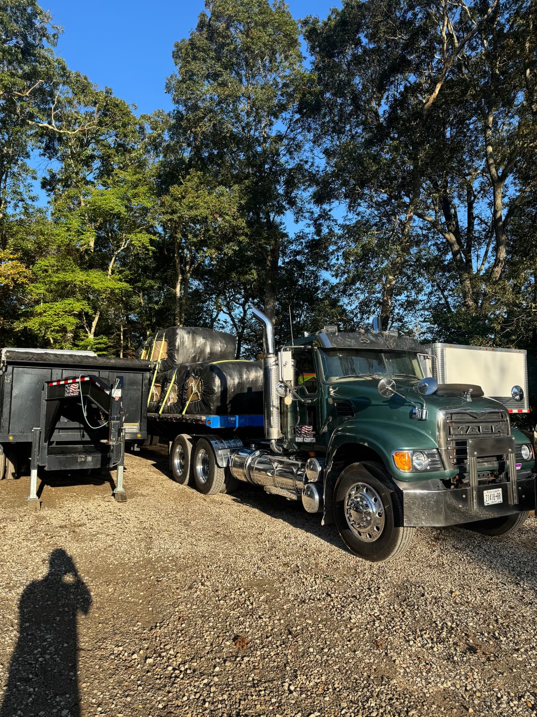 Green heavy-duty truck parked beside black trailer in wooded area. Clear blue sky above.